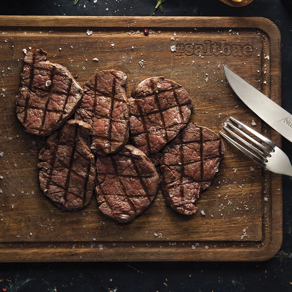 A beautifully presented wooden board featuring grilled tenderloin slices, arranged neatly with visible grill marks, lightly seasoned with coarse salt, accompanied by a fork and knife engraved with the Nusret logo.