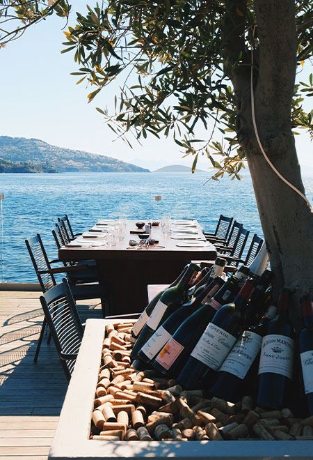 Seaside dining at Nusr-Et Yalıkavak, featuring a wine display under an olive tree with scenic views of the Aegean Sea, perfect for a luxurious meal.