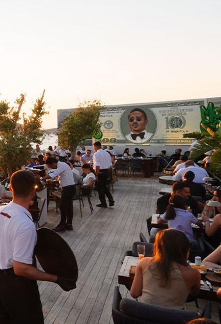 Lively dining area at Nusr-Et Yalıkavak, featuring a Salt Bae mural and outdoor seating with beautiful views of the sea at sunset